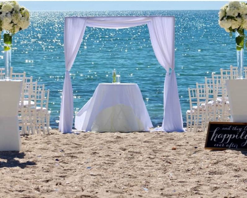 Ceremony setup on the beach: a white draped arch facing the sea with white chairs on both sides
